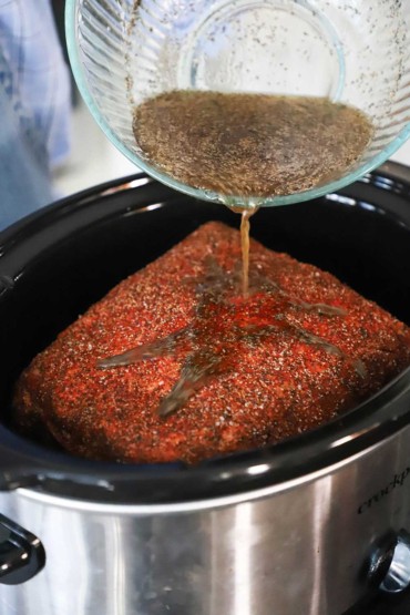 A person pouring a liquid mop mixture over a Boston pork butt that is seasoned and sitting in a slow cooker.