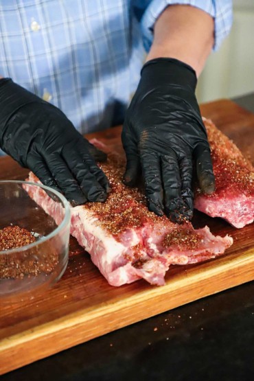A person wearing protective latex gloves pressing a pork rub into two slab of baby back ribs on a large cutting board.