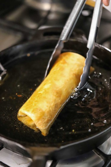 A person using metal tongs to hold a flour tortilla that has been wrapped like a beef burrito and is being deep-fried in a skillet filled with hot oil.