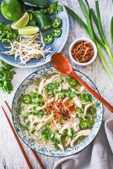 An overhead view of a bowl of chicken pho garnished with cilantro, peppers, and fried shallots, sitting next a platter of additional garnishes, including lime wedges and mung bean sprouts.