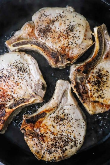 An overhead view of four seared bone-in pork chops in a large cast-iron skillet.