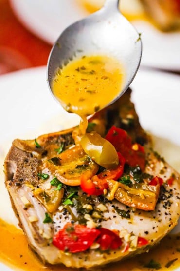A straight-on view of a large spoon transferring a savory brown sauce over the top of a seared pork chop that is covered with Italian peppers, garlic, and herbs.