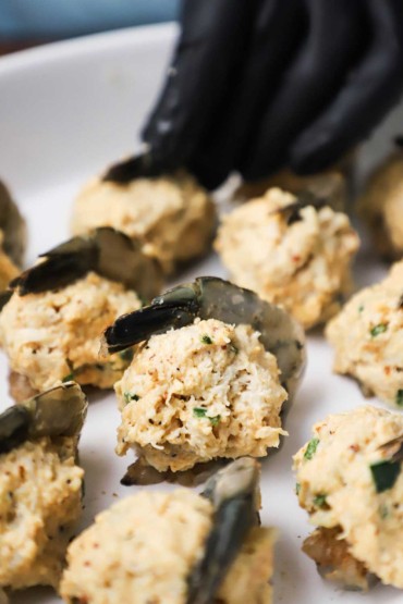 A close-up view of a shrimp in a baking dish that is sitting upright and its tail is wrapped around an uncooked crab mixture.