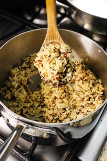 A large wooden spoon being used to lift up a large helping of a cooked wild rice medley over a saucepan filled with the rice.