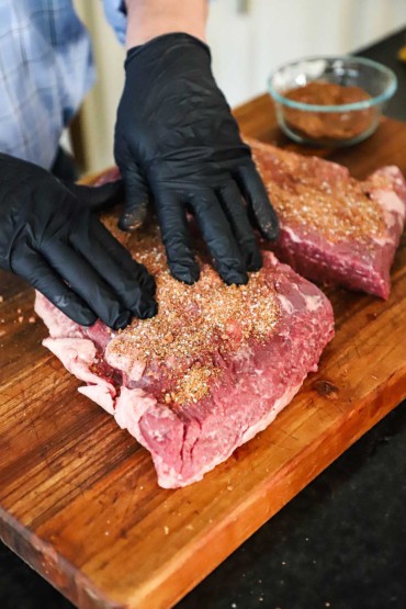 A person wearing black cooking gloves pressing a barbecue rub into a brisket flat that has been cut into two pieces.