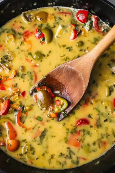 An overhead view of a large black cast-iron skillet that is filled with a light brown sauce filled with sautéed peppers, garlic, and herbs, with a wooden spoon inserted in the middle of it all.