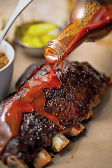A person pouring homemade barbecue sauce from a clear glass bottle onto a rack of slow cooker baby ribs that have been fully cooked.