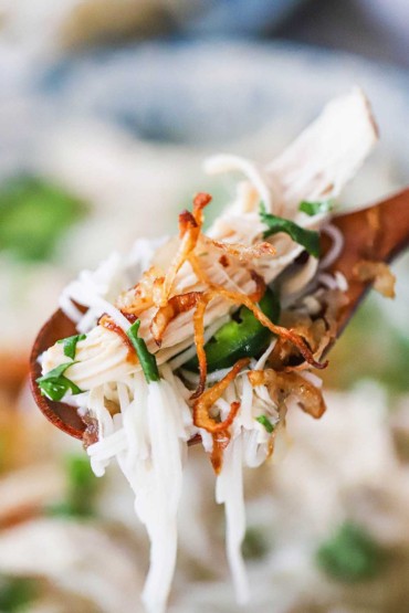 A wooden spoon being used to lift up a helping of chicken pho that is loaded with thin noodles, shredded chicken, herbs, and fried shallots.