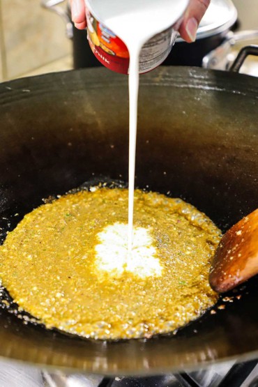 Unsweetened coconut cream being poured from a glass milk jug into a wok filled with green curry sauce.