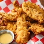 A straight-on view of a basket that is lined with a red-checkered piece of wax paper and is filled with a pile of crispy chicken tenders and a small container of honey mustard dipping sauce.