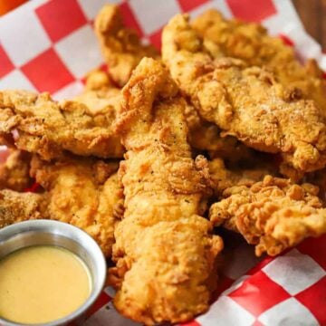 A straight-on view of a basket that is lined with a red-checkered piece of wax paper and is filled with a pile of crispy chicken tenders and a small container of honey mustard dipping sauce.