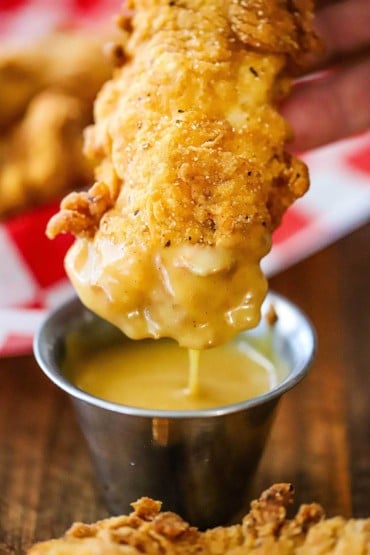 A close-up view of a crispy chicken tender that has been dipped in a small container of homemade honey mustard dipping sauce a is dripping from the bottom of the tender.