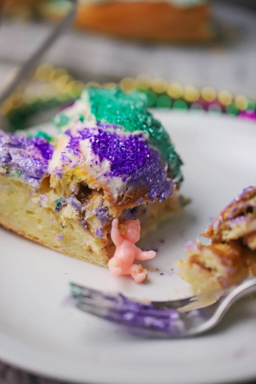 A close-up view of a piece of Mardi Gars King Cake that is half eaten and a small baby is resting against the side of the piece of cake.