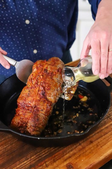 A person pouring white wine from a small glass carafe under a stuffed flank steak that is being held up in a large cast-iron skillet.
