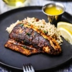 A straight-on view of a blackened redfish filet resting against a bed rice pilaf with two lemon wedges and a small vessel of melted butter nearby.