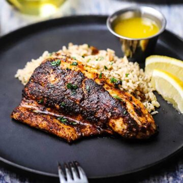 A straight-on view of a blackened redfish filet resting against a bed rice pilaf with two lemon wedges and a small vessel of melted butter nearby.