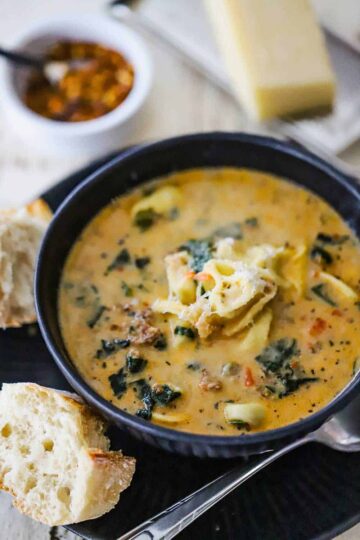 A close-up view of a blue soup bowl filled with creamy tortellini soup topped with grated parmesan cheese with two chunks of Italian bread sitting nearby.