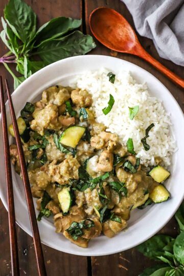An overhead view of a shallow white bowl filled on one side with Thai green curry chicken and the other side with steamed Jasmine rice and topped with chopped Thai basil.
