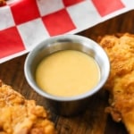 A close-up view of a small silver condiment vessel filled with homemade honey mustard dipping sauce surround by chicken tender.