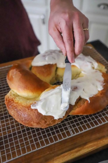 A person using a flat-edge spatula to smear white icing over the top of a braided cake.