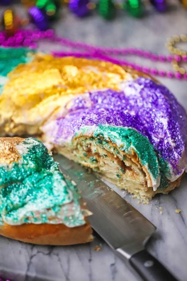 A straight-on view of a colorful Mardi Gras King cake that has a slice missing where the cinnamon filling is visible and a large knife is next to the cake.