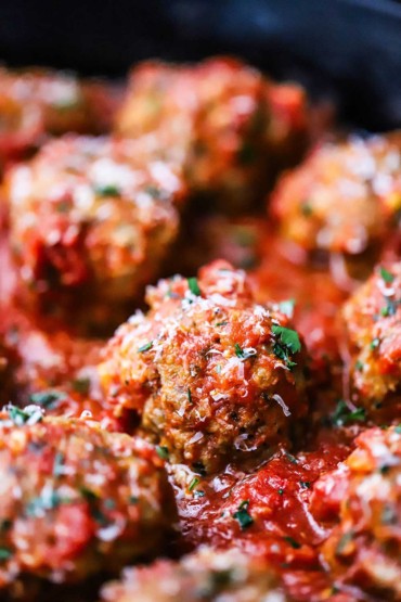 A close-up view of Italian meatballs in a marinara sauce topped with shaved parmesan and chopped parsley.
