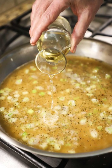 A person pouring white wine from a small carafe into a large skillet filled with simmering broth with chopped celery and uncooked long grain rice.