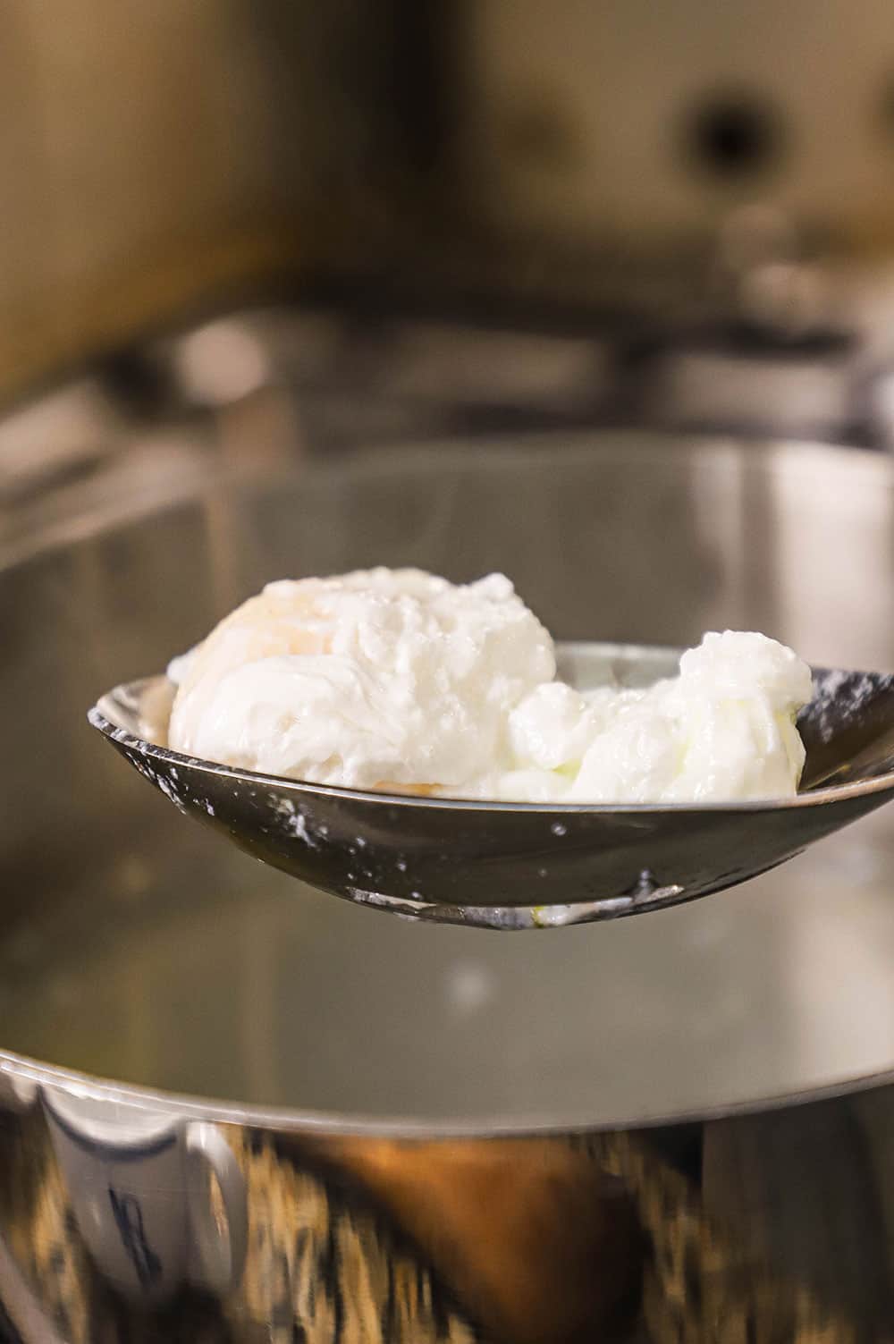 A silver slotted spoon lifting up holding two fluffy poached eggs over a pan full of hot steaming water.