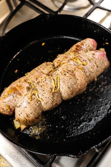 An overhead view of a stuffed flank that has been rolled and tied with twine and is being seared in a cast-iron skillet.