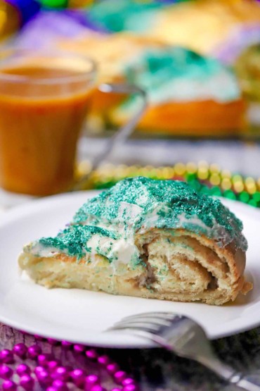 A close-up view of a slice of Mardi Gras King cake with glitter green icing on top and a circular cinnamon filling is visible.