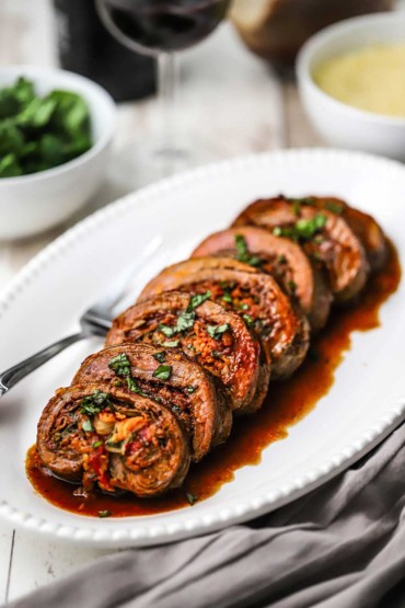 A straight-on view of a white oval platter filled with slices of stuffed flank steak that has been covered in a wine reduction sauce.