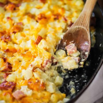 A close-up view of a wooden spoon that is inserted into a cheesy ham and potato casserole in a black baking dish with a portion of the casserole missing.