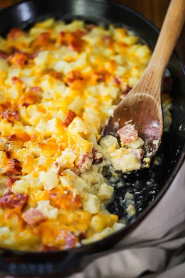 A close-up view of a wooden spoon that is inserted into a cheesy ham and potato casserole in a black baking dish with a portion of the casserole missing.