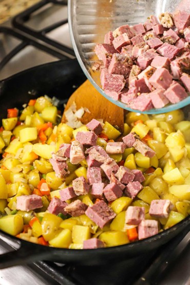 A person transferring cubed corned beef from a glass bowl into a skillet filled with cubes of yellow potatoes, and chopped red bell pepper.