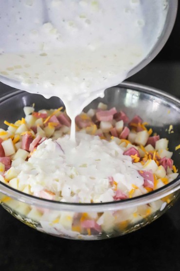 A creamy béchamel sauce being poured from a skillet into a large glass bowl filled with cubed potatoes, shredded cheddar cheese, and chunks of ham.
