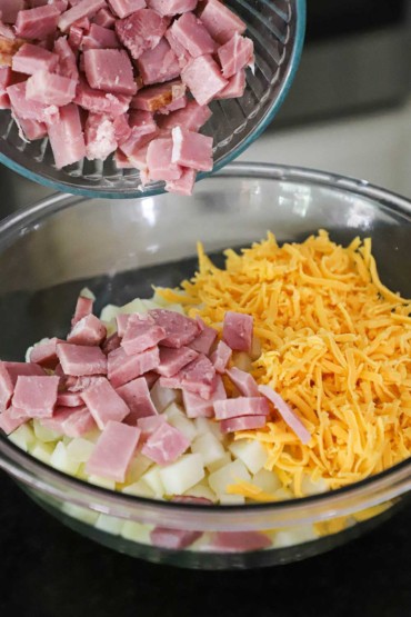 Chunks of ham being dumped from one glass bowl into a large glass bowl that is filled with a pile of cubed potatoes and topped with shredded cheddar cheese.