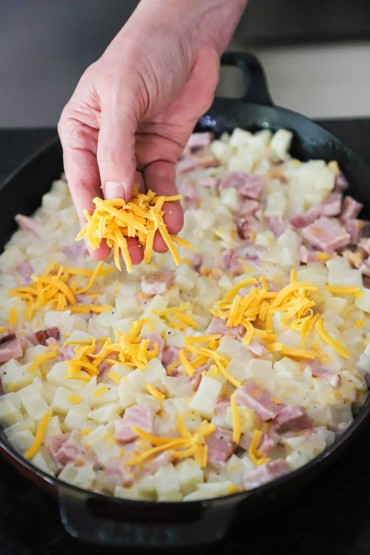 Person sprinkling shredded cheddar cheese over the top of an unbaked ham and potato casserole in an oval black baking dish.