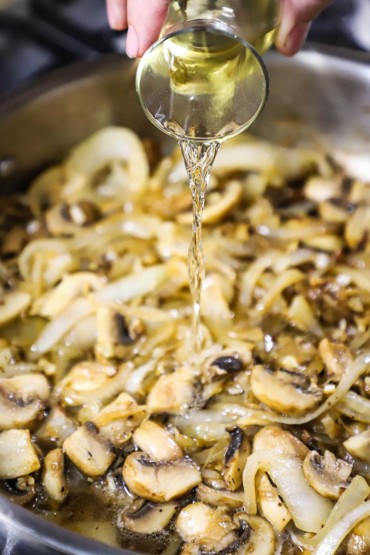 A person pouring white wine from a small carafe into a large silver skillet filled with sautéed onion slivers and sliced mushrooms.