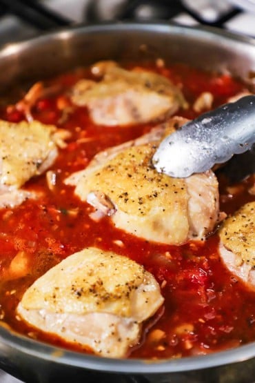 A metal pair of tongs being used to nestle a seared chicken thigh into a skillet filled with a simmering tomato sauce and several other chicken thighs.