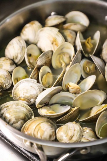 A close-up view of littleneck clams that have opened their shells and are simmering in white wine in a large stainless steel skillet.