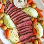 An overhead view of a large white oval platter that is filled with sliced corned beef flanked by sautéed cabbage, carrots, and baby yellow potatoes.