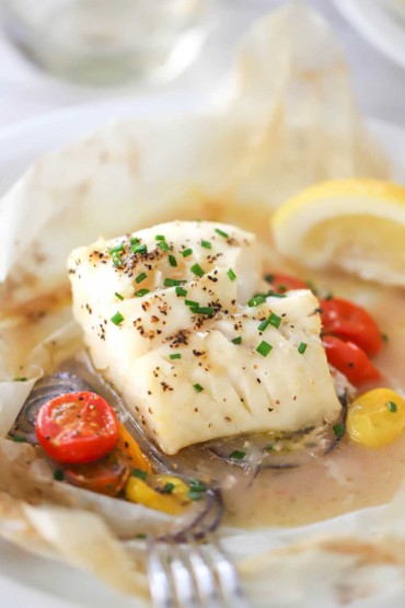 A close-up view of a steamed fillet of cod that has been cooked in a packet of parchment paper along with white wine, herbs, cherry tomatoes, and red onion.