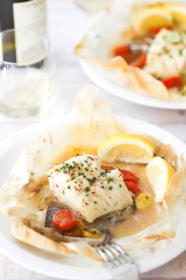 A straight-on view of two dinner plates that are holding servings of fish en papillote with a lemon wedge off to the side.