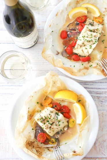 An overhead view of two dinner plates that are holding servings of fish en papillote with lemon wedges on the side of the plate and a bottle of wine nearby.