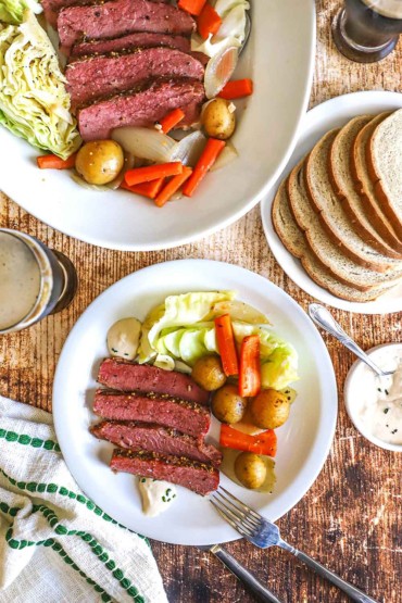 An overhead view of a dinner plate filled with corned beef and cabbage next to a plate of sliced rye bread and a large platter of more corned beef, cabbage, potatoes, and carrots.