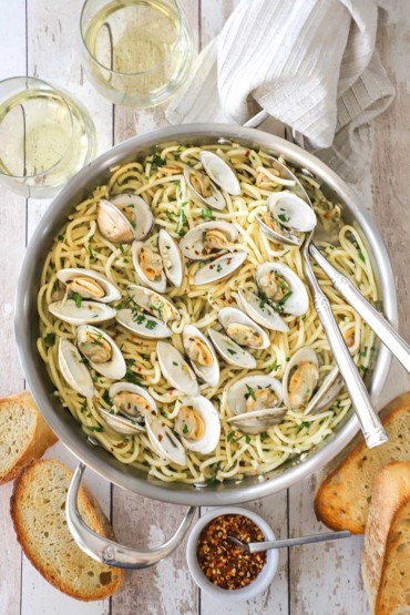 An overhead view of a large stainless steel skillet filled with spaghetti all vongole with opened clams resting on top of the cooked pasta and two serving utensils inserted into the side of the dish.