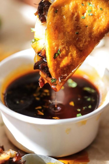 A close-up view of a beef birria taco that is being dipped into a small bowl of beef consommé.