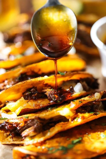 A straight on view of a person using a metal spoon to drizzle consommé over a row of beef birria tacos that are arranged side by side on a board.