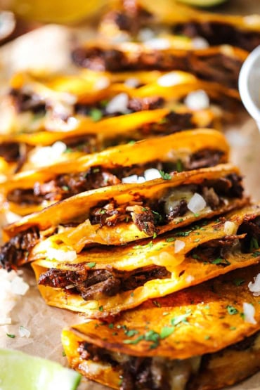 A straight-on view of a row of beef birria tacos that are in a row each one resting on top of the taco next to it and they are garnish with chopped onion and cilantro.