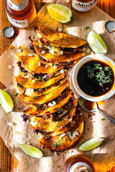 An overhead view of a row of beef birria tacos that are on laying on their sides next to a white dipping bowl filled with beef consommé and surrounded by lime wedges and Mexican bottled beer.